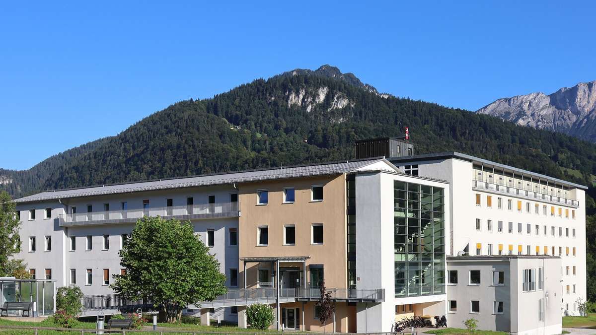A modern building with multiple floors, a glass wall, and several windows. Trees and a bench are in the foreground, with mountains in the background.