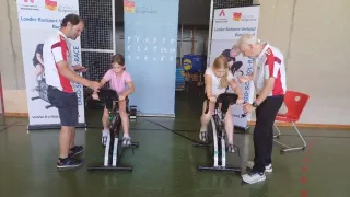 Two girls ride exercise bikes in a gym, assisted by two men. Banners in German are displayed behind them.