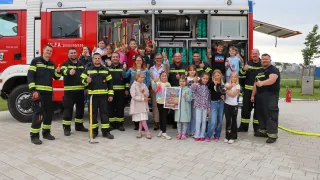 Firefighters and children stand in front of a fire engine, smiling and posing for a photo. A girl holds a colorful poster.