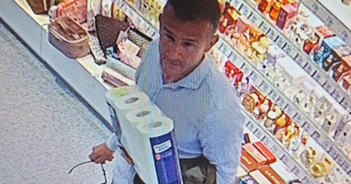 A man is inside a convenience store, holding a toilet paper package in his hand. Behind him, there are shelves filled with snacks.