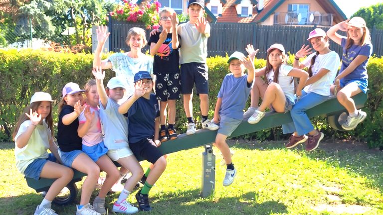 A group of children and adults sitting on a seesaw in a garden, all smiling and waving. Behind them, there are bushes, a fence, and a house with a balcony.