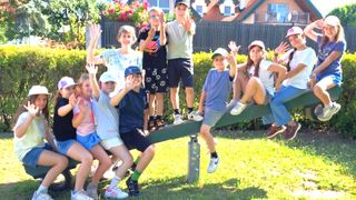 A group of children and adults sitting on a seesaw in a garden, all smiling and waving. Behind them, there are bushes, a fence, and a house with a balcony.