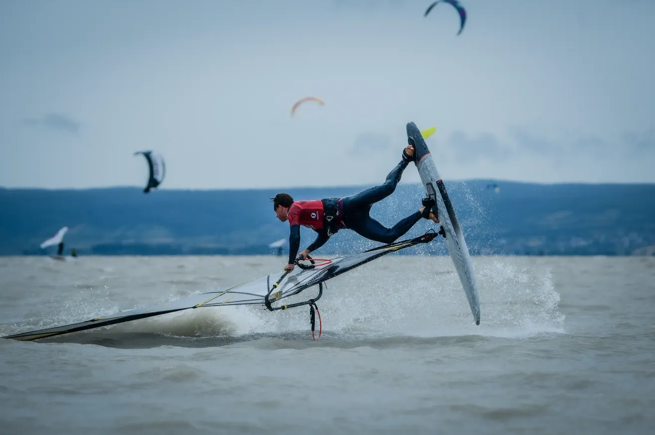 A windsurfer in a red suit performs a stunt on a board while water splashes around.