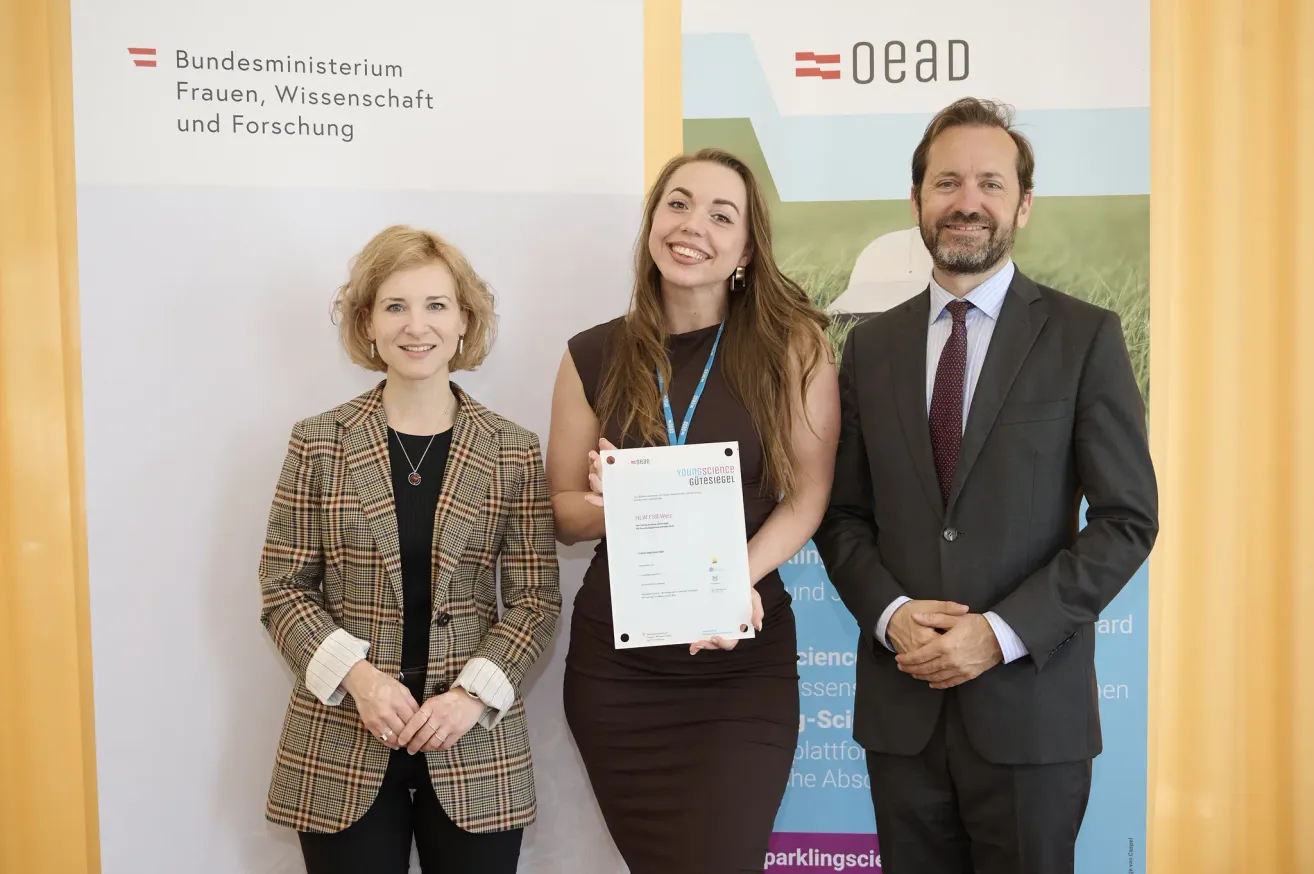 Three people stand in front of a backdrop with the word 'OEAD'. A woman holds a certificate and smiles. The other two people, a man and woman, look at her.