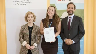 Three people stand in front of a backdrop with the word 'OEAD'. A woman holds a certificate and smiles. The other two people, a man and woman, look at her.