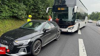 A black car and a white double-decker bus are parked on the roadside. Two people in safety gear are standing next to the bus.