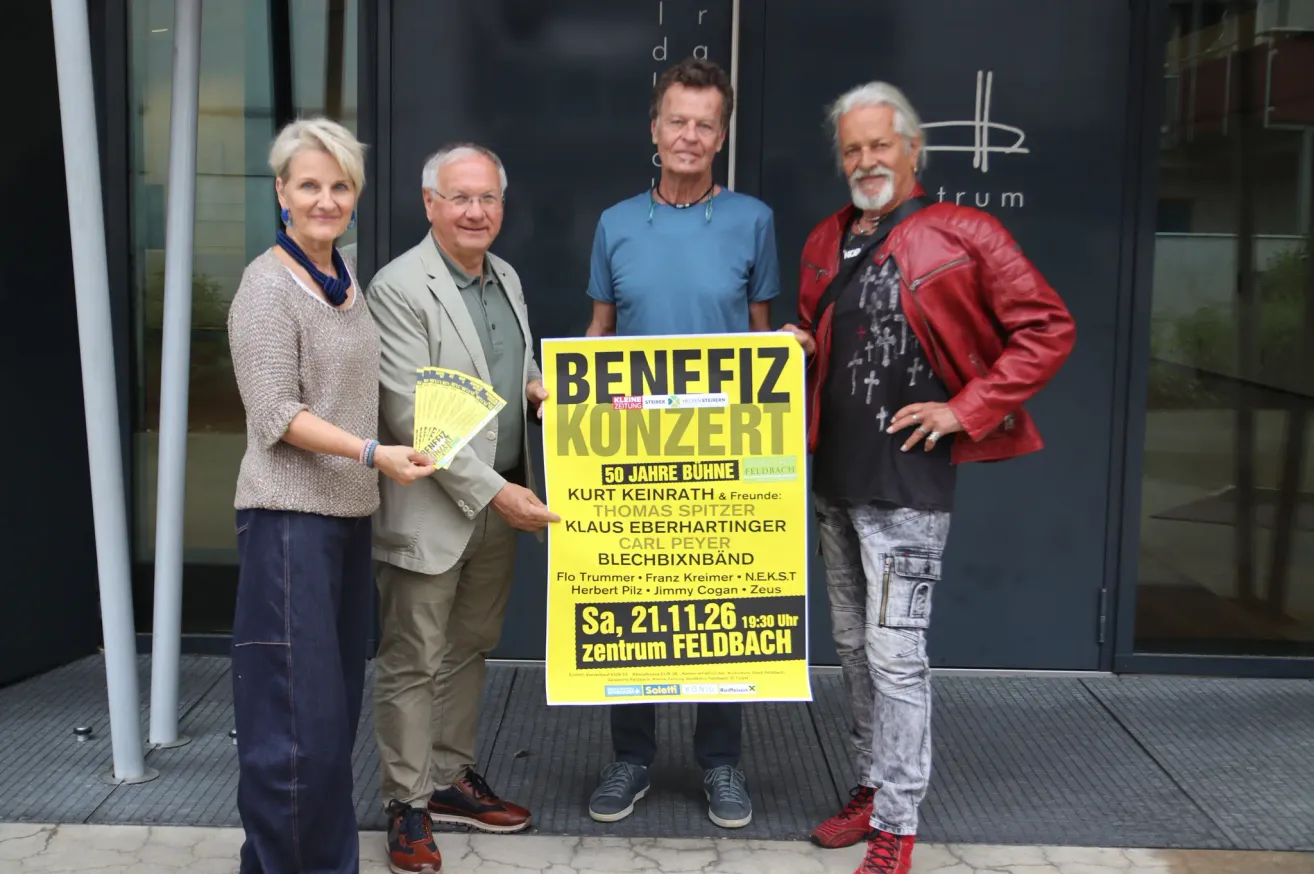 Four people stand in front of a building, holding a poster that says Benefiz Konzert. They are dressed casually and appear to be posing for a photo. The poster has text and images on it.