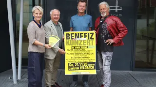 Four people stand in front of a building, holding a poster that says Benefiz Konzert. They are dressed casually and appear to be posing for a photo. The poster has text and images on it.