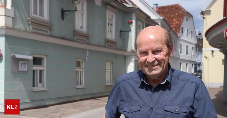 Ein älterer Mann in einem blauen Hemd lächelt vor einem traditionellen europäischen Gebäude.