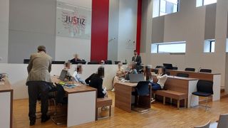 A court hearing with the word Justiz on the wall. People are seated, and one man stands with papers. Several laptops and books are on the tables.