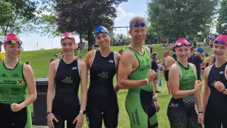 Three people dressed in swim gear stand together on a grass field. Two women wear black and one man wears green. They all wear caps and goggles. Other people are in the background, some swimming.