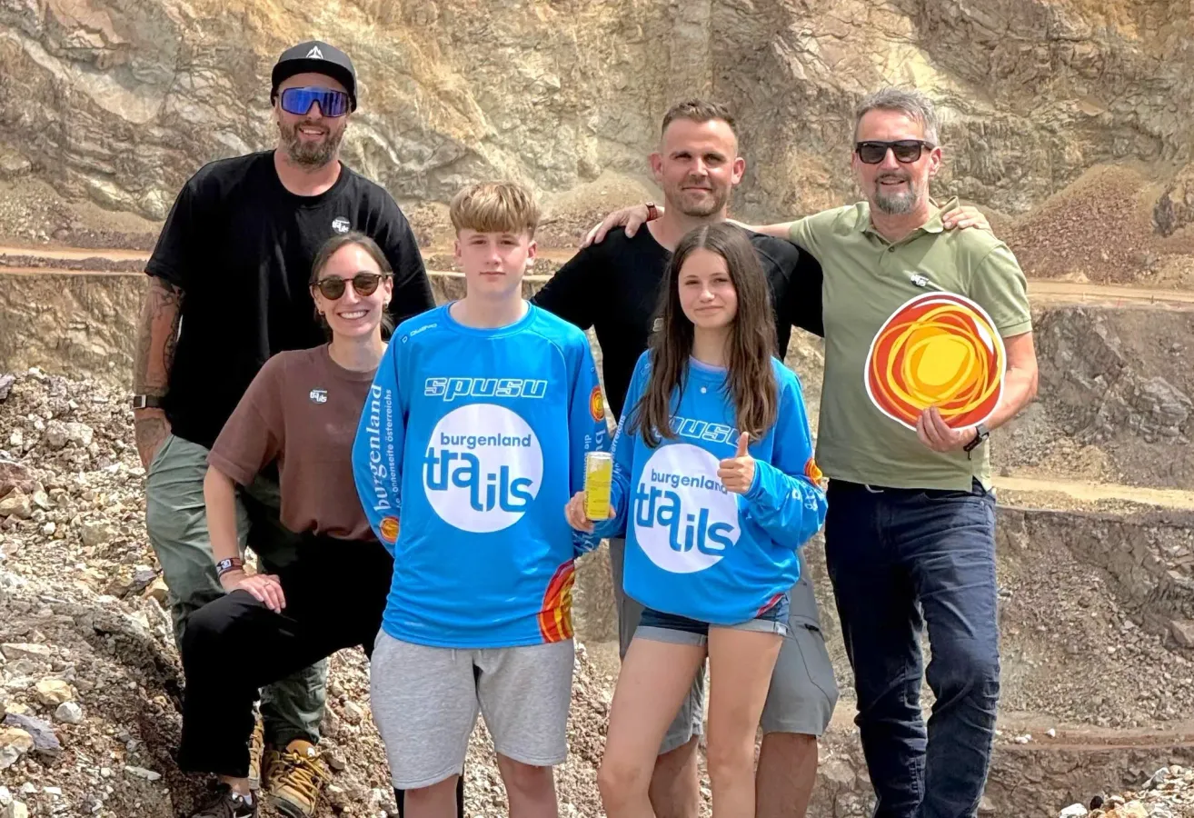 A group of adults and children pose for a photo in front of a mountain. They wear matching blue Burgenland Trails shirts.
