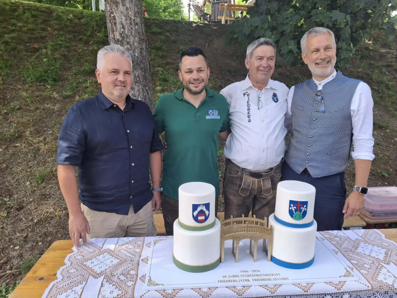 Four men stand around a table, smiling. One holds a cake, which has two levels, each with a city emblem on it. The cake has the words 'Friedberg' and '60 Jahre Stadtfreundschaft' written on it.