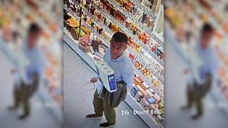 A man in a supermarket carrying two rolls of toilet paper. He wears a blue shirt and green pants.