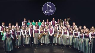 A group of musicians dressed in traditional attire, holding various instruments, pose for a photo on stage.
