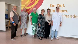 Five individuals, including two women in glasses, are standing together in front of a wall with a vibrant sun graphic and text.
