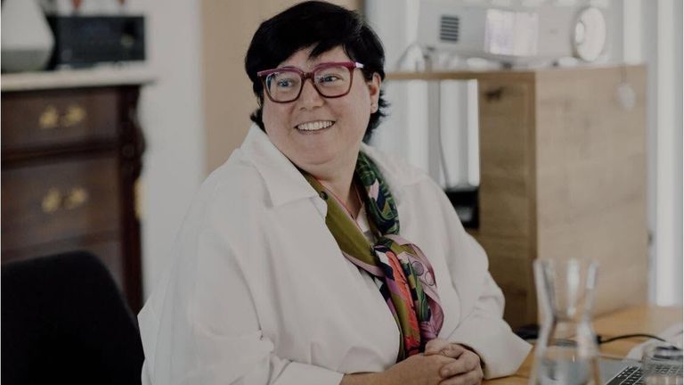 A woman with short black hair wearing glasses and a white lab coat, smiling and posing for a photo in an indoor setting.