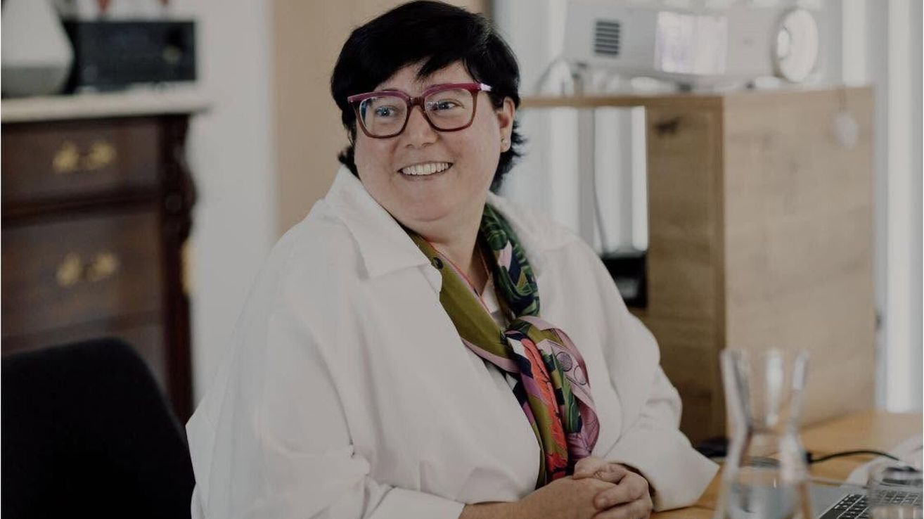 A woman with short black hair wearing glasses and a white lab coat, smiling and posing for a photo in an indoor setting.