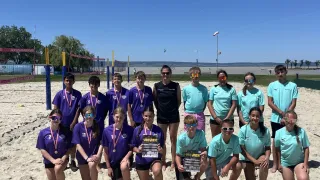 A beach volleyball team poses for a photo on the sand, wearing matching purple and teal jerseys. They have medals around their necks and hold a trophy. The sky is clear.