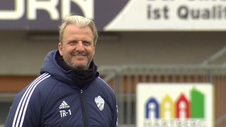 A smiling man wearing a blue Adidas jacket with 'TR-1' on it stands in front of a blurry fence and a sign with white letters.