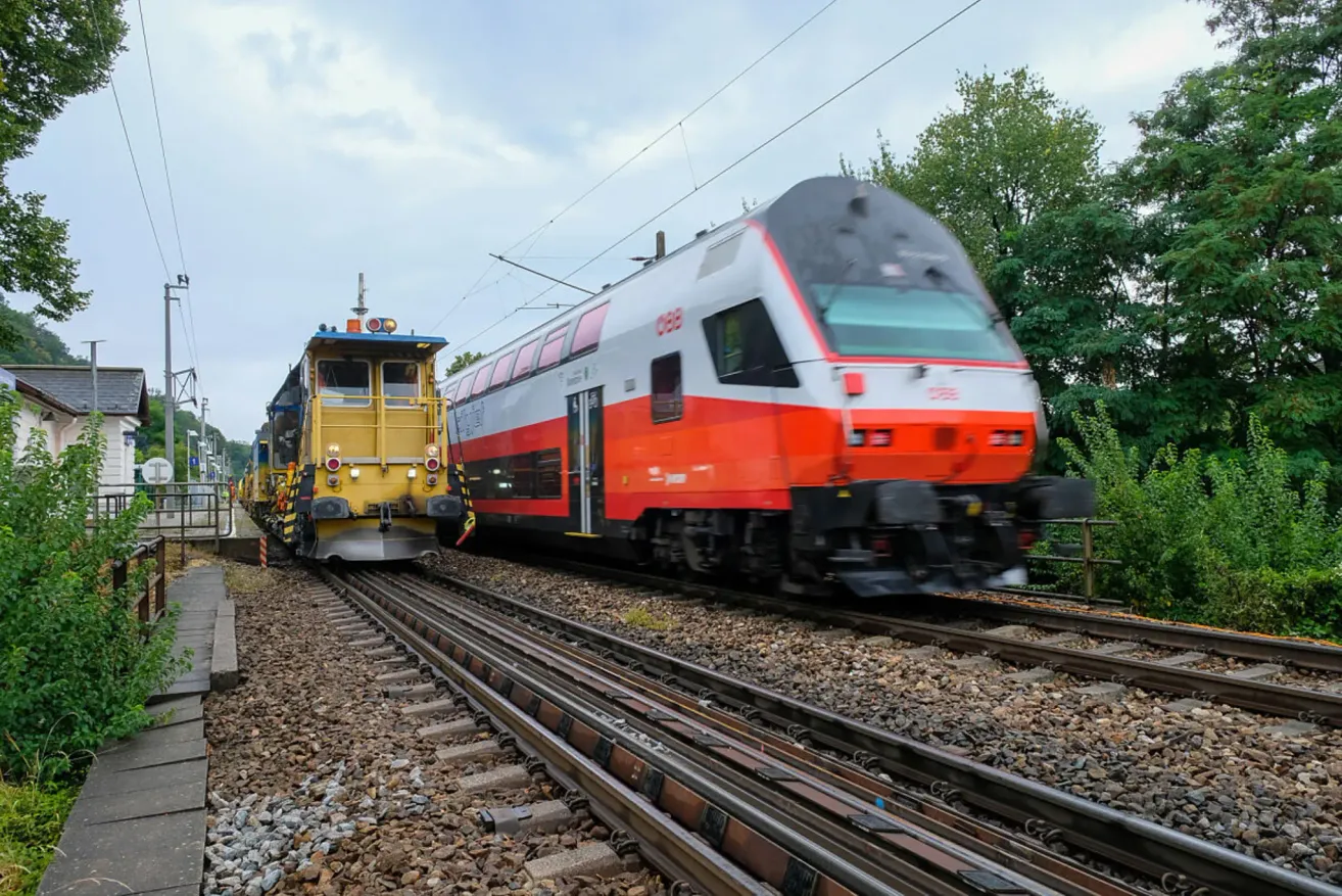 Two trains are traveling on a railroad track, one in yellow and the other in orange and white. Trees are on the side of the tracks.