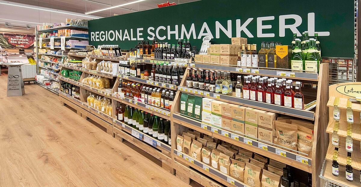A store with a variety of regional snacks and drinks. The shelves are filled with bottles of liquor, bottles of syrup, and bags of nuts. A sign at the top reads Regionale Schmankerl.
