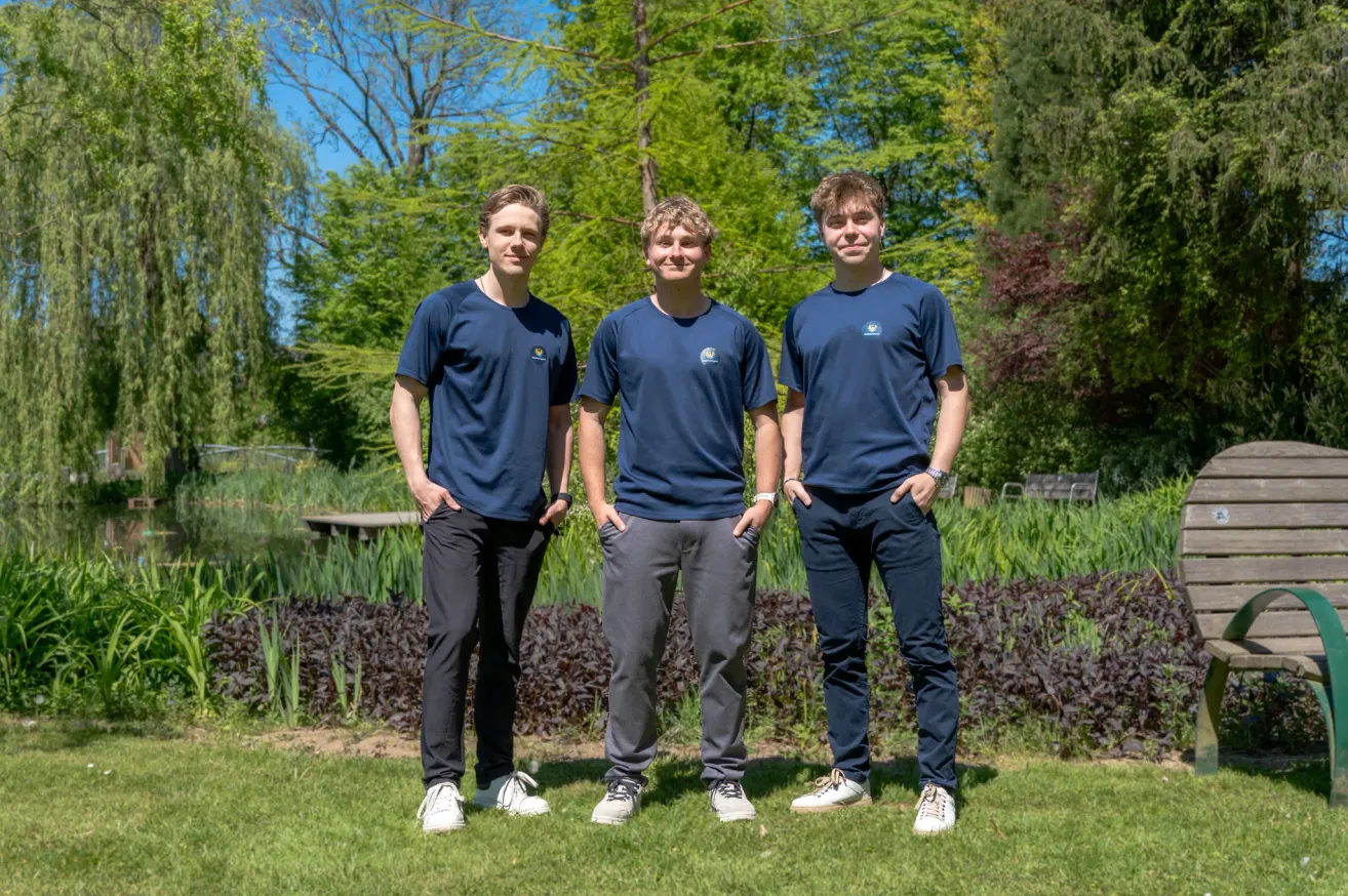 Three young men in matching navy blue shirts and gray pants stand together on a lawn. They are smiling and appear to be posing for a photo. Behind them, a garden with green trees and plants can be seen.