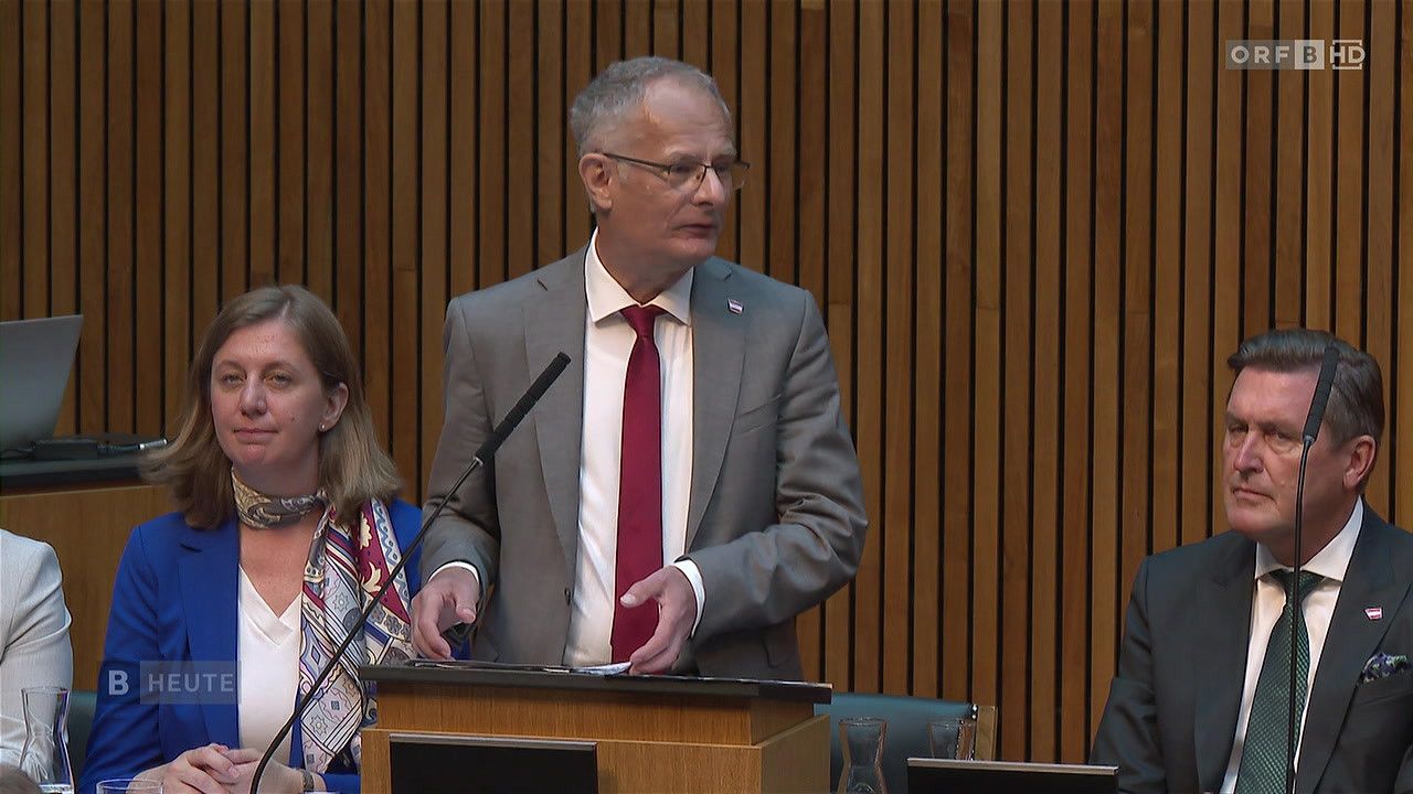 Ein Mann in einem grauen Anzug, roter Krawatte und Brille steht an einem Podium mit einem Mikrofon. Eine Frau steht zu seiner Linken und eine Holzwand befindet sich hinter ihm.