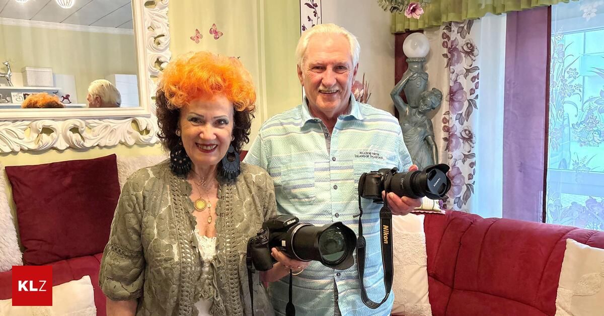 Ein älterer Mann und eine ältere Frau stehen in einem Wohnzimmer, lächeln und posieren mit zwei großen Nikon-Kameras. Hinter ihnen befindet sich ein Blumenvorhang und ein Spiegel mit cremefarbenem Rahmen.