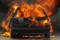 A black car is engulfed in flames on a street, with the rear end most affected.