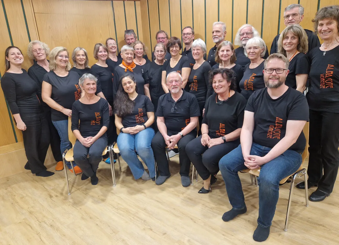 A choir group in matching black t-shirts with the word 'Aldeote' on them, posing for a photo in a room.