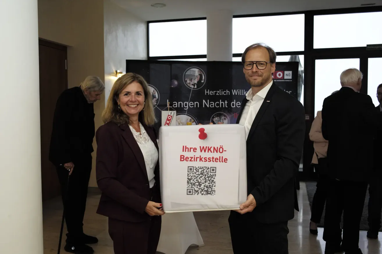 A man and woman stand indoors, smiling and holding a white bag with a QR code, 'Ihre WKNö-Bezirksstelle' written on it. Behind them, a banner with images and text is visible.