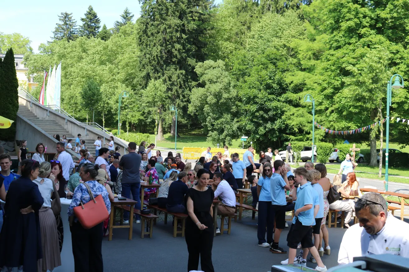 A large group of people are gathered in an outdoor setting, possibly for an event. They are sitting and standing around wooden tables and benches, with some wearing glasses and hats. There are trees and streetlights in the background.