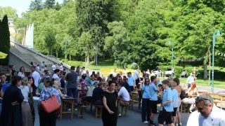 A large group of people are gathered in an outdoor setting, possibly for an event. They are sitting and standing around wooden tables and benches, with some wearing glasses and hats. There are trees and streetlights in the background.