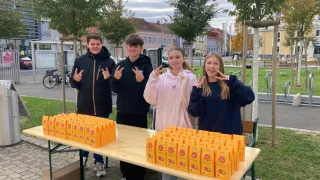 Four individuals stand behind a table filled with orange bags, smiling and posing for a photo. The background shows trees, buildings, and a pathway.