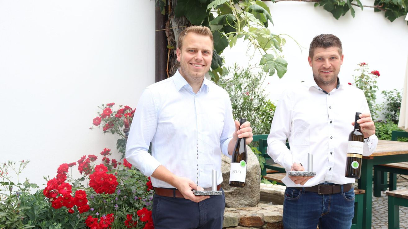 Two men stand in front of a garden, holding awards and wine bottles. The man on the left wears a white shirt and blue pants, while the man on the right wears a white shirt and jeans.