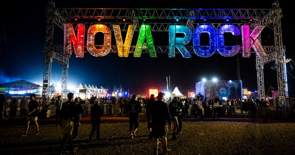 Eingang zum Nova Rock Festival mit großen bunten Buchstaben, Menschen gehen am Abend mit Zelten und Lichtern im Hintergrund.
