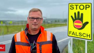 A man in an orange safety vest is standing on the side of a road. Behind him, a yellow sign reads STOP.