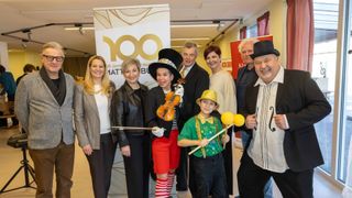 A group of adults and children dressed in costumes pose for a photo in a hall, with a woman holding a violin and a boy holding drumsticks.