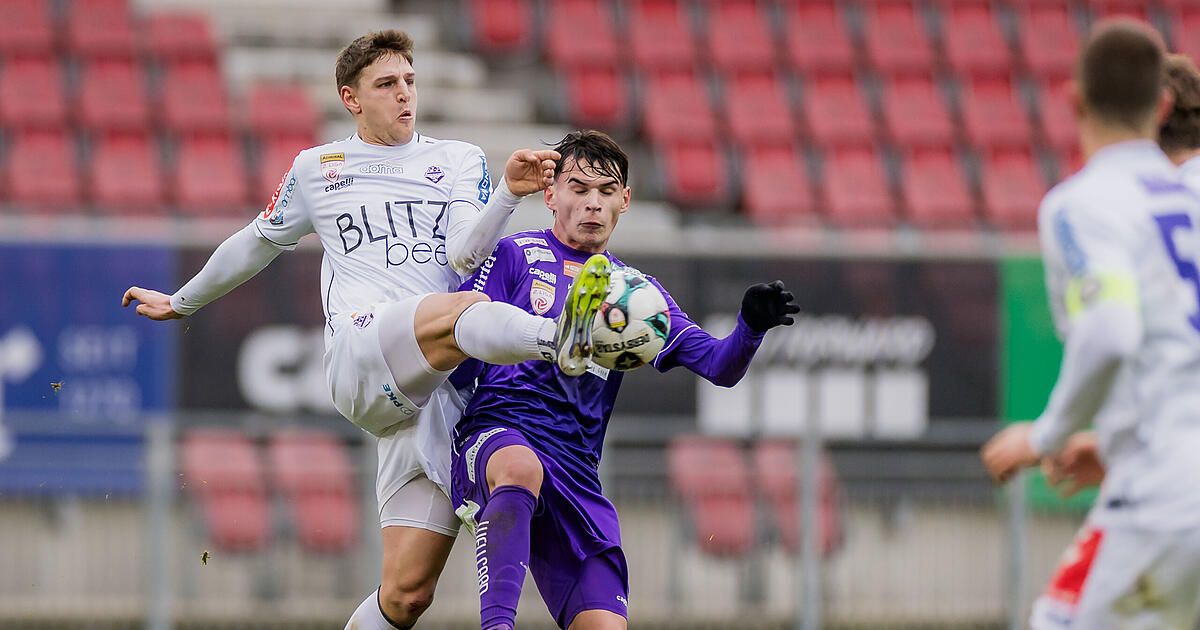 Two soccer players, one in white and the other in purple, are playing on a field. One is jumping to kick the ball, and the other is trying to block. Behind them are empty red chairs.