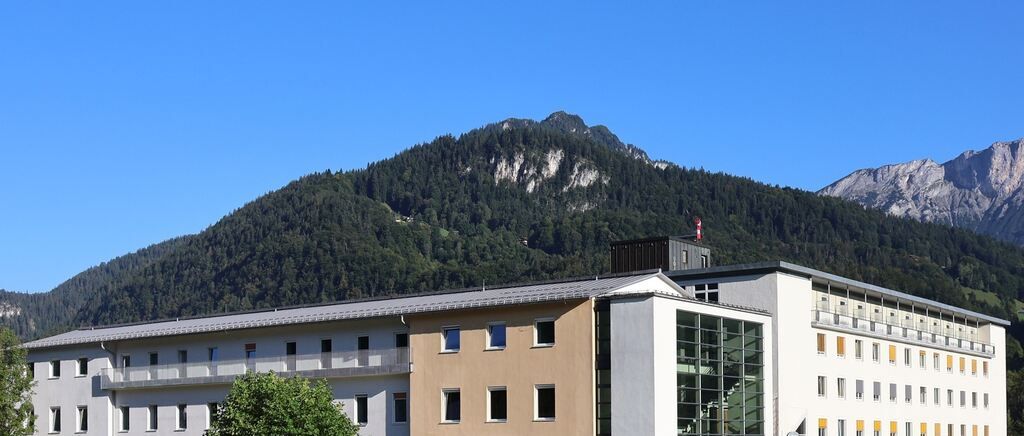 A building with multiple windows and a balcony stands before a mountain covered in trees. The sky is clear and blue.