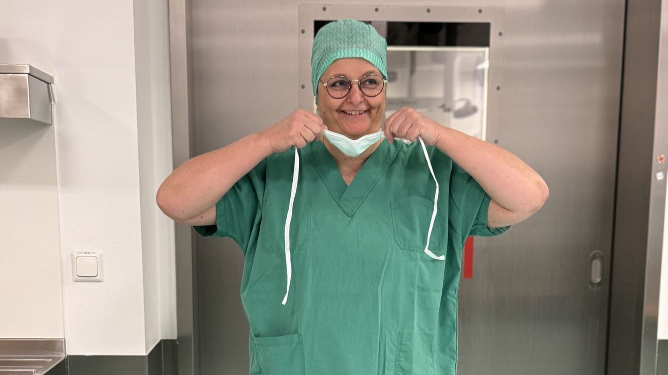 Eine lächelnde Frau in grüner Arbeitskleidung justiert ihre Maske in einem Krankenhauskorridor. Sie trägt eine Brille und eine grüne Kappe. Hinter ihr befindet sich eine Metalltür und ein Glasfenster.