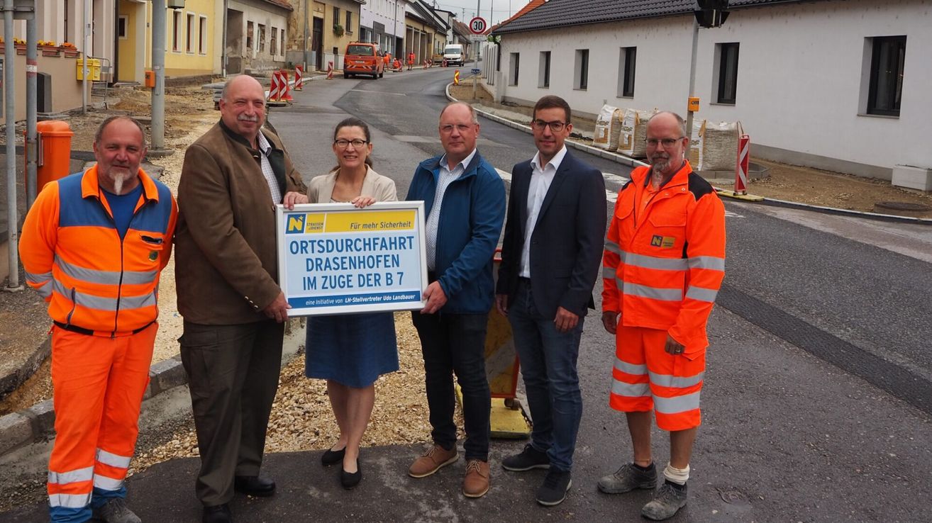 Fünf Personen stehen auf einer Straße und halten ein Schild für Ortsdurchfahrt Drasenhofen im Zuge der B 7, eine lokale Initiative. Sie tragen verschiedene Arbeits- und formelle Kleidung. Hinter ihnen ist eine Baustelle zu sehen mit Verkehrskegeln und einem 30 km/h Geschwindigkeitsschild.