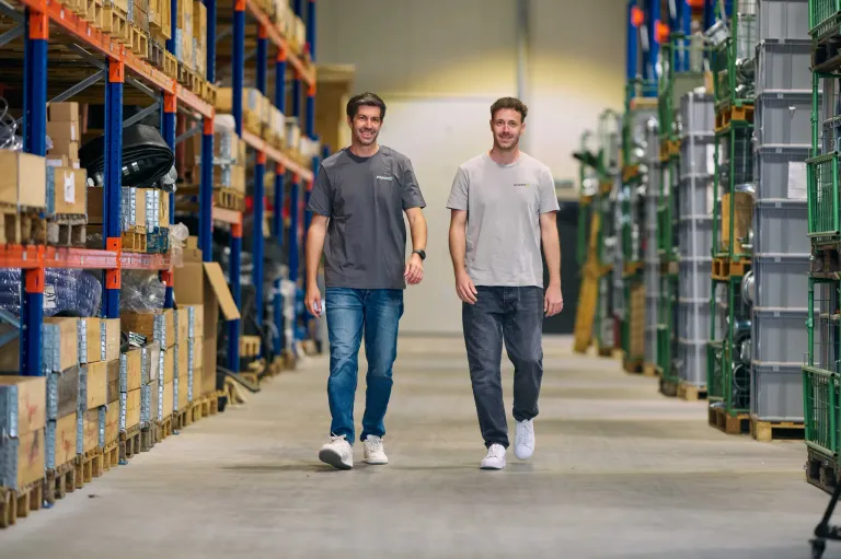 Zwei Männer in lässiger Kleidung gehen durch ein Lager, lächeln und scheinen für ein Foto zu posieren. Hinter ihnen sind Regale mit verschiedenen Artikeln gefüllt, und der Boden ist sauber und gut beleuchtet.