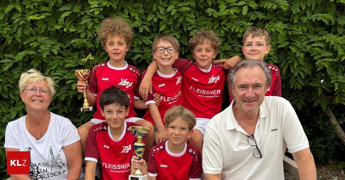 Eine Gruppe junger Jungen in roten Uniformen posiert mit ihren Trophäen und einem Mann in einem weißen Poloshirt, lächelnd für ein Foto vor einem grünen Hintergrund.