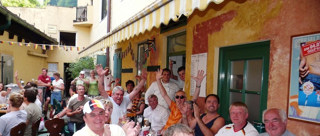A group of people celebrate outside a yellow-walled building with a striped awning. They wave and smile, some seated with drinks. The building features a green door and a whiteboard with writing.
