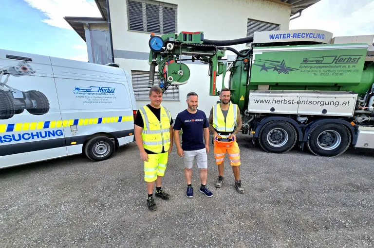 Drei Männer in Sicherheitswesten stehen vor einem Gebäude, mit einem großen grünen Recycling-Lkw im Hintergrund.