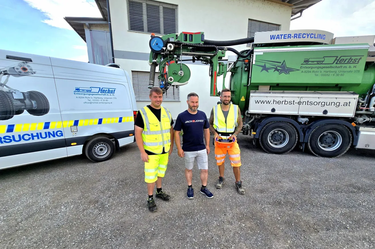Drei Männer in Sicherheitswesten stehen vor einem Gebäude, mit einem großen grünen Recycling-Lkw im Hintergrund.