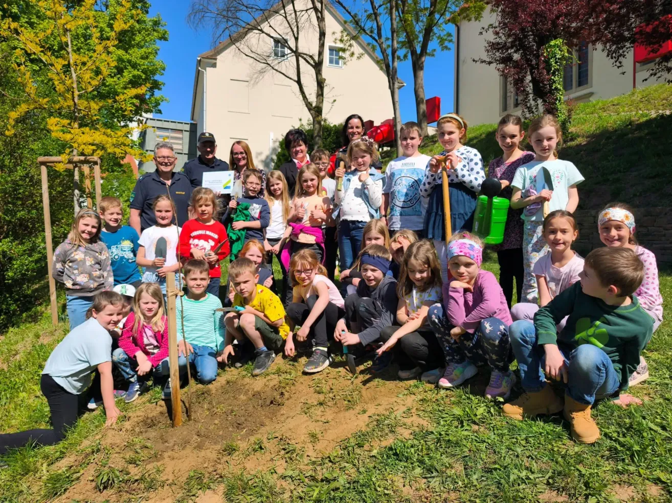 A group of children and adults are planting a tree in a garden. Some children are holding shovels and others are standing and watching. In the background are houses and trees.