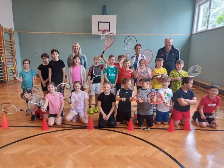 Eine Gruppe von Kindern und zwei Erwachsenen posiert mit Tennisschlägern in einem Gymnasium für ein Foto.